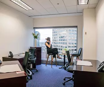 Servcorp serviced office featuring a modern, professional workspace with multiple desks, ergonomic chairs, and a cityscape view through a large window, where a woman is seated on a high stool talking on the phone.