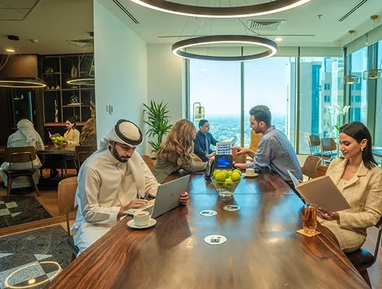 A vibrant coworking space at Sahab Tower, Kuwait, where professionals are engaged in work and conversation around a shared table, surrounded by natural light and city views.